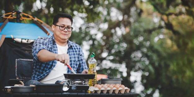 Ritratto di uomo viaggiatore asiatico occhiali bistecca di maiale frittura barbecue in padella o pentola per arrostire in un campeggio concetto di stile di vita campeggio viaggio cucina all'aperto