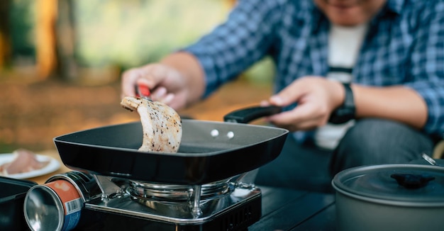 Ritratto di uomo viaggiatore asiatico occhiali bistecca di maiale frittura barbecue in padella o pentola per arrostire in un campeggio concetto di stile di vita campeggio viaggio cucina all'aperto