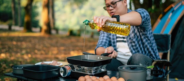 Ritratto di uomo viaggiatore asiatico occhiali versando olio di semi di girasole in una padella Cucina all'aperto concetto di stile di vita in campeggio in viaggio