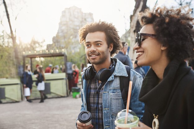 Ritratto di vista laterale dell'affascinante ragazzo afro-americano con taglio di capelli afro che osserva da parte mentre cammina con la sua ragazza nel parco, beve caffè e si gode una serata calda