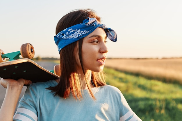 Ritratto di vista laterale di bella donna pensosa che indossa una maglietta casual blu e un'elegante fascia per capelli, distogliendo lo sguardo con sguardo premuroso, tenendo lo skateboard sulle spalle.