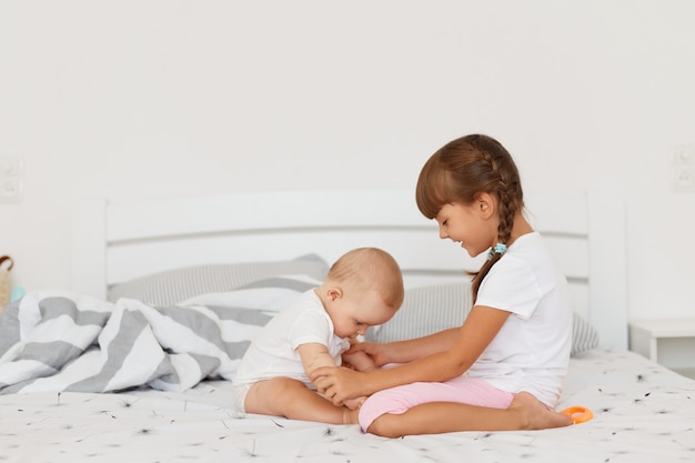 Ritratto di vista laterale di ragazze carine e felici sedute sul letto in camera da letto luminosa, bambini che indossano abiti bianchi che giocano insieme a casa, trascorrono del tempo insieme, infanzia felice.