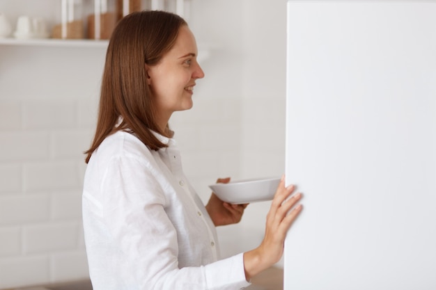 Ritratto di vista laterale di una femmina dai capelli scuri positiva che indossa una camicia bianca, apre il frigorifero, trova cibo per colazione o cena, guardando sorridente all'interno del frigorifero.