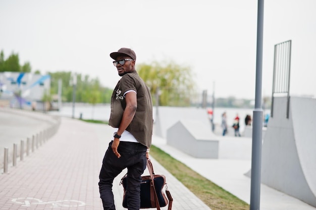 Ritratto di vista posteriore di camminare alla moda uomo afroamericano indossare occhiali da sole e berretto con borsa all'aperto Street fashion uomo nero