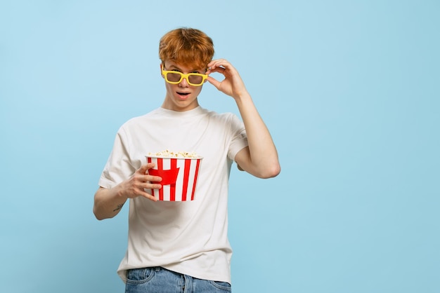 Ritratto di yong ragazzo emotivo in occhiali 3D con cesto di popcorn in posa isolato su sfondo blu dello studio Tempo del film