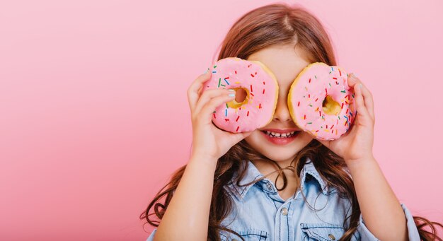 Ritratto eccitato gioiosa giovane ragazza graziosa in camicia blu che esprime positività, divertendosi alla macchina fotografica con ciambelle sugli occhi isolati su sfondo rosa. Infanzia felice con gustosi dessert. Posiziona il testo