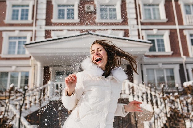 Ritratto eccitato ragazza allegra che si diverte con la neve in una mattina di sole ghiacciata sulla strada. Emozioni vere, risate ad occhi chiusi, momenti luminosi, atmosfera natalizia, vacanze invernali