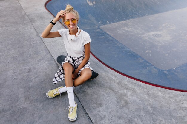 Ritratto esterno di donna emotiva che si siede sullo skateboard. Donna abbronzata agghiacciante in calzini bianchi e scarpe sportive che trascorrono del tempo in skate park.