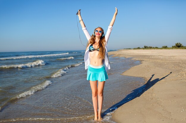 Ritratto estivo all'aperto di ragazza carina felice che si diverte e impazzisce vicino all'oceano, colori solari e atmosfera positiva, abbigliamento da spiaggia alla moda luminoso, ascolto di musica in cuffia