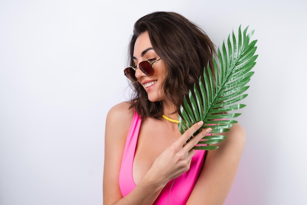 Ritratto estivo di giovane donna in costume da bagno rosa collana da spiaggia giallo brillante con foglia di palma monstera e occhiali da sole