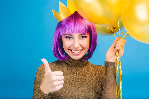 Ritratto felice che celebra momenti di gioiosa giovane donna con palloncini dorati sorridente. Abito di lusso, capelli viola tagliati, corona da principessa, umore allegro.