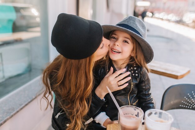 Ritratto felice famiglia amorevole insieme. Madre e figlia seduti in un caffè della città e giocando e abbracciando. Bambina felice guardando la telecamera, madre che bacia la figlia sulla guancia.