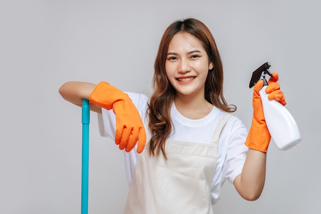 Ritratto felice giovane bella donna in grembiule e guanti di gomma in possesso di un flacone spray che si prepara a pulire, sorridere e guardare la macchina fotografica, copia spazio