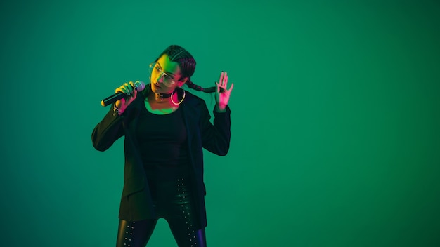 Ritratto femminile caucasico della cantante isolato sulla parete verde alla luce al neon. Bellissimo modello femminile in abbigliamento nero con microfono. Concetto di emozioni umane, espressione facciale, pubblicità, musica, arte.