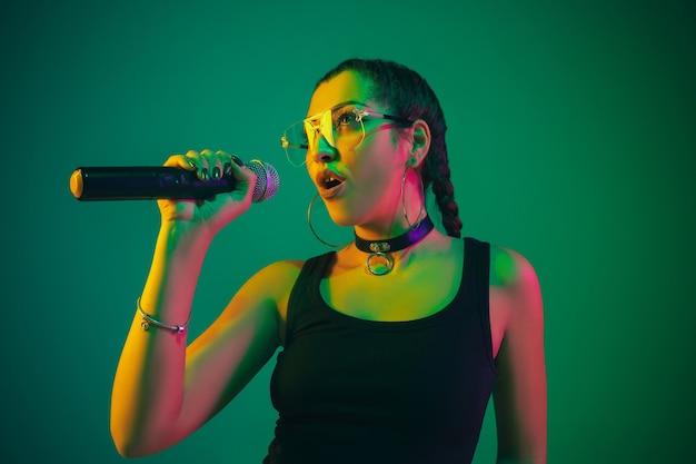 Ritratto femminile caucasico della cantante isolato sulla parete verde alla luce al neon. Bellissimo modello femminile in abbigliamento nero con microfono.