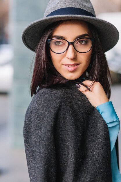 Ritratto gioiosa giovane donna alla moda con capelli castani in vetri neri che camminano sulla strada in città. Cappotto grigio, cappello, stile di vita di lusso, aspetto elegante