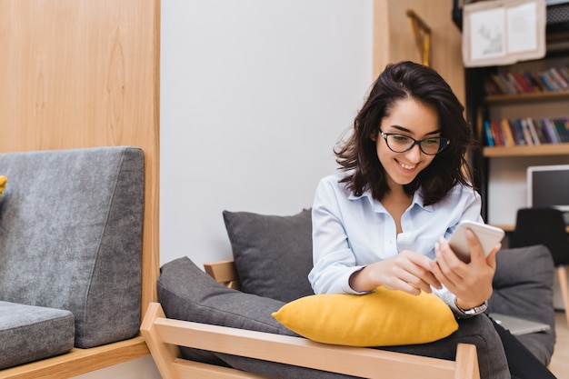 Ritratto giovane donna bruna gioiosa in vetri neri agghiacciante sul divano in appartamento moderno. Usare il telefono, mandare SMS, sorridere, umore allegro.