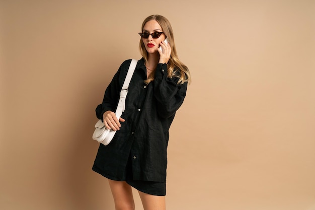 Ritratto in studio alla moda di donna alla moda che parla dal suo lavoro di influencer su smartphone indossando un abito di lino nero