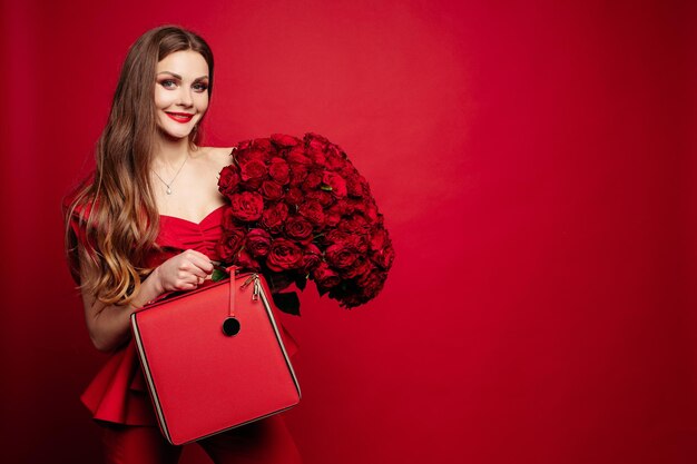 Ritratto in studio alla moda di giovane donna alla moda con lunghi capelli castani in abito rosso costoso con borsa in pelle rossa e mazzo di rose Sta sorridendo alla telecamera Labbra rosse Sfondo rosso