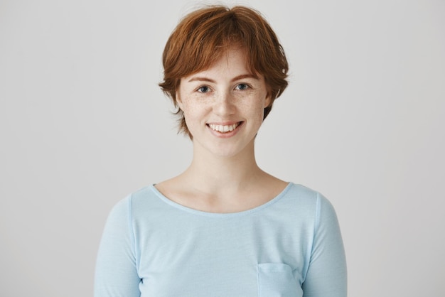 Ritratto in studio di carismatica donna caucasica attraente con colore rosso naturale dei capelli e lentiggini che sorride allegramente mentre in piedi su sfondo grigio in posa per la pubblicità