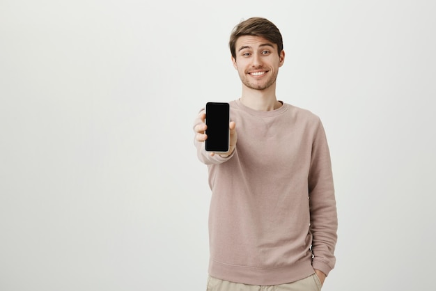 Ritratto in studio di felice attraente modello maschio europeo che mostra smartphone mentre sorride e tiene una mano in tasca
