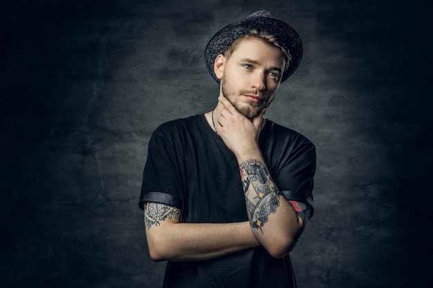 Ritratto in studio di maschio premuroso con braccia tatuate incrociate, vestito con una maglietta nera e un cappello di tweed.