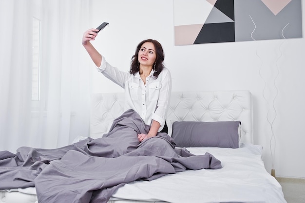 Ritratto in studio di ragazza bruna in camicetta bianca sdraiata sul letto e utilizzando il telefono cellulare
