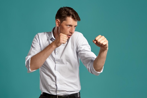 Ritratto in studio di un bel giovane uomo d'affari in una classica camicia bianca che si comporta come se stesse combattendo mentre posa su uno sfondo blu. Taglio di capelli alla moda. Concetto di emozioni sincere. Copia spazio.