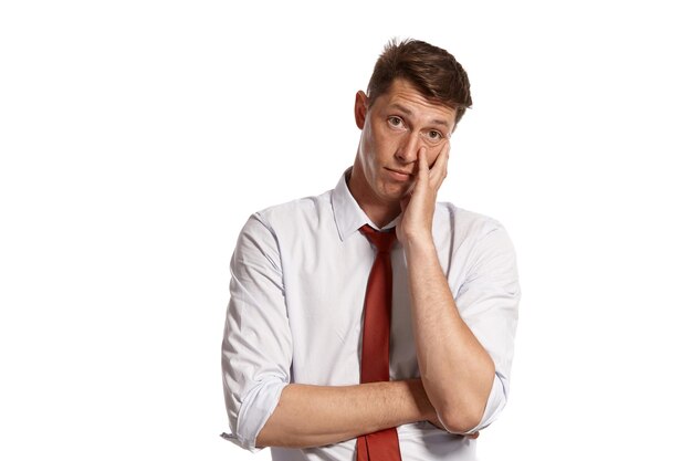 Ritratto in studio di un giovane lunatico in una classica camicia bianca e cravatta rossa che sembra stanco e in posa isolato su sfondo bianco. Taglio di capelli alla moda. Concetto di emozioni sincere. Copia spazio.