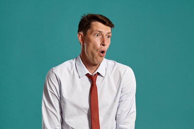 Ritratto in studio di un giovane uomo carismatico in una classica camicia bianca e cravatta rossa che sembra meravigliato mentre posa su uno sfondo blu. Taglio di capelli alla moda. Concetto di emozioni sincere. Copia spazio.