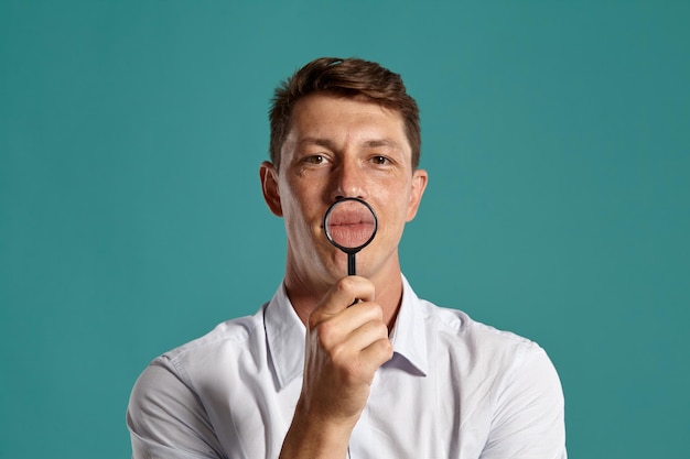 Ritratto in studio di un giovane uomo d'affari atletico in una classica camicia bianca che mostra le sue labbra attraverso una lente d'ingrandimento mentre posa su uno sfondo blu. Taglio di capelli alla moda. Concetto di emozioni sincere. Copia spazio