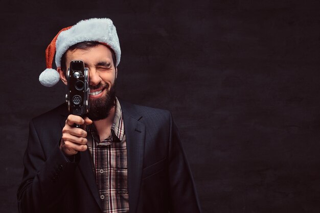 Ritratto in studio di un uomo barbuto sorridente che indossa un abito classico e un cappello da Babbo Natale che tiene una fotocamera retrò, facendo una foto. Isolato su uno sfondo scuro.