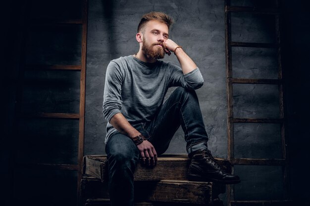 Ritratto in studio di uomo urbano barbuto in jeans si siede su una scatola di legno con scale vintage sullo sfondo.