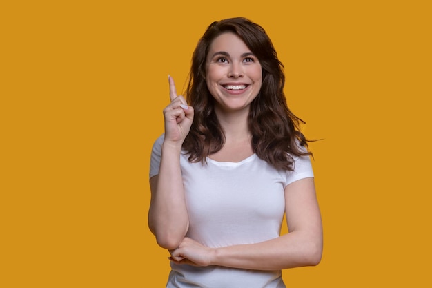 Ritratto in vita di una ragazza caucasica dai capelli scuri sorridente e compiaciuta che punta verso l'alto con l'indice sollevato