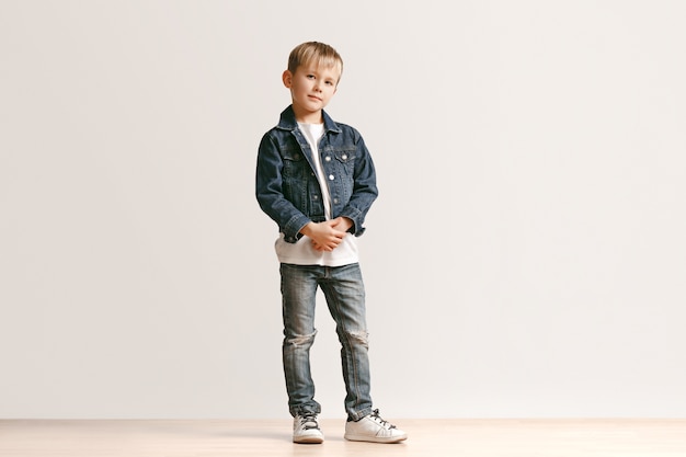 Ritratto integrale del ragazzino sveglio in vestiti di jeans alla moda e sorridente, in piedi sul bianco. Concetto di moda per bambini