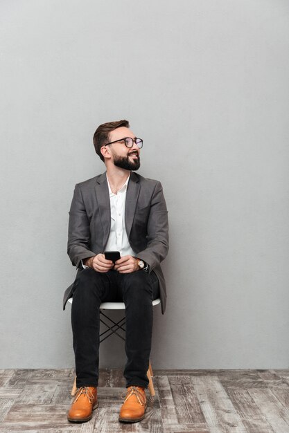 Ritratto integrale dell'uomo rilassato nella seduta casuale sulla sedia in ufficio che guarda da parte con lo smartphone in mani, isolato sopra grey