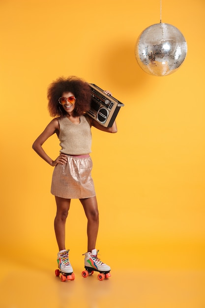 Ritratto integrale della donna africana allegra della discoteca con la mano sulla sua vita che indossa in retro vestiti che stanno sui pattini di rullo, tenendo boombox