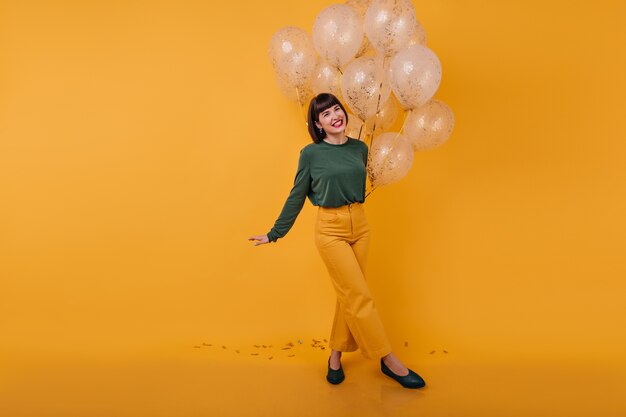 Ritratto integrale della donna che ride in piedi con le gambe incrociate. Tiro al coperto di ragazza di compleanno romantico ballando con palloncini dorati.