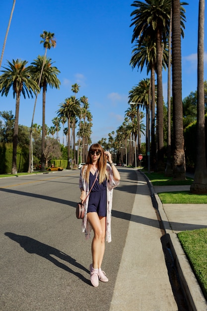 Ritratto integrale della donna sorridente alla moda che cammina sulla strada esotica vicino all'hotel in una giornata calda e soleggiata. Trascorre le sue vacanze a Los Angeles