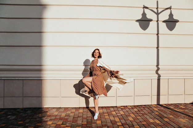 Ritratto integrale della ragazza attiva che balla nella giornata di sole. Foto all'aperto di una donna riccia disinvolta che scherza per strada.