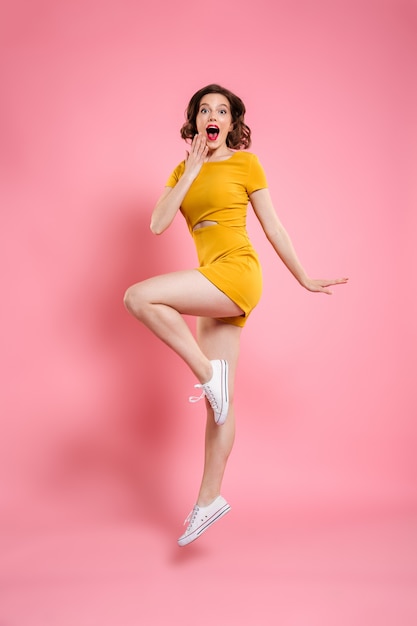 Ritratto integrale della ragazza graziosa uscita felice in vestito giallo elegante mentre saltando sopra il rosa