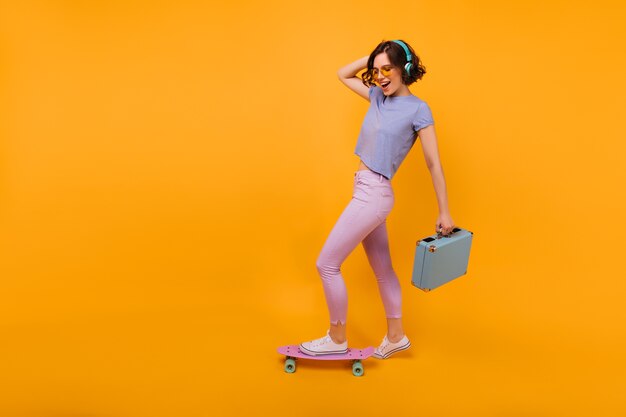 Ritratto integrale della ragazza positiva con la posa blu della valigia. Modello femminile riccio giocondo in piedi sul longboard e sorridente.