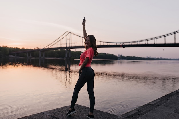 Ritratto integrale della ragazza sportiva che gode della vista sul fiume in serata. Ritratto esterno di splendida signora sorridente facendo fitness sulla natura.