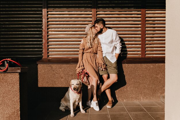 Ritratto integrale delle coppie che baciano su fondo di legno. Ragazza in abito a pois e il suo ragazzo barbuto alla moda in posa con il cane.