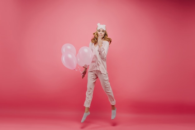 Ritratto integrale di donna entusiasta in pigiama che salta con palloncini al mattino. Felice ragazza di compleanno con i capelli ricci che scherza sul muro rosa brillante.