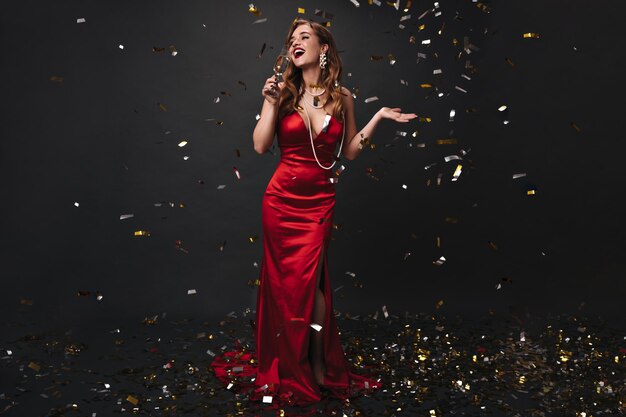 Ritratto integrale di donna in abito rosso che beve champagne su sfondo nero
