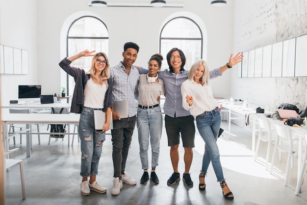 Ritratto integrale di impiegato femminile sottile in jeans che stanno con le gambe attraversate vicino al collega asiatico. Foto dell'interno dello studente africano alto e della donna europea felice con il computer portatile.