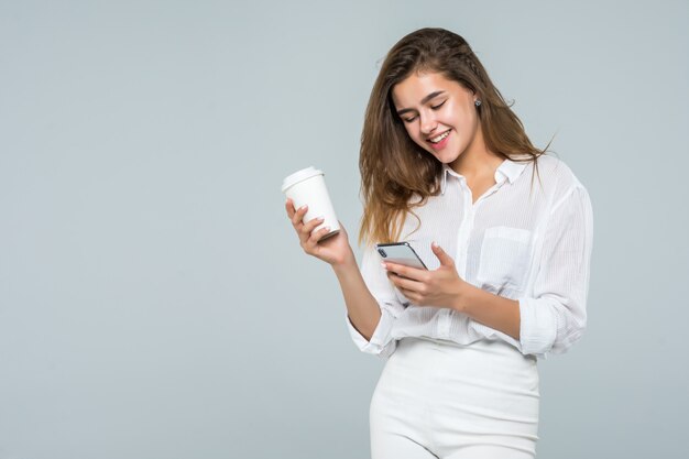 Ritratto integrale di una ragazza sorridente felice che per mezzo del telefono cellulare mentre stando e tenendo la tazza di caffè sopra fondo bianco