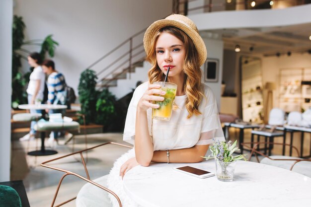 Ritratto interno di una giovane donna adorabile che beve succo di frutta gustoso con menta al tavolo con fiori su di esso. Foto di una ragazza elegante in abito bianco che si rilassa al bar con persone sullo sfondo.