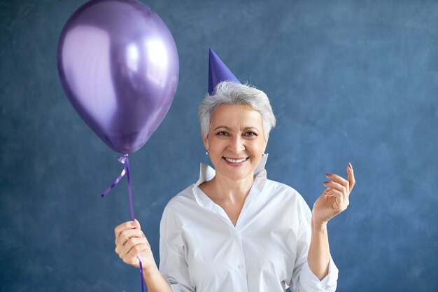 Ritratto isolato di riuscita imprenditrice matura con corti capelli grigi in posa al muro bianco con palloncino di elio